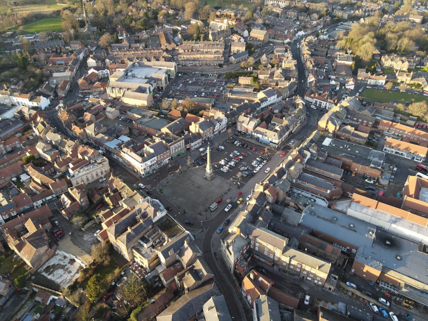 An aerial aerial view of Harlington High Street showing a small town centre with a mix of residential and commercial buildings, narrow streets, and parked cars. In the foreground, a town square is visible with a central monument or war memorial, surrounded by a paved area with vehicles parked around it. The image captures the dense layout of buildings, rooftops with tiled and flat surfaces, and streets extending outward towards greener areas with trees. The lighting suggests late afternoon or early evening, casting shadows across the scene. This overview highlights the urban setting typical for home relocation and furniture transport services, with visible roadways and parking spaces indicating potential parking opportunities for a removal vehicle or van, as provided by companies like Man With a Van Harlington during furniture moving and packing processes in the context of house removals.