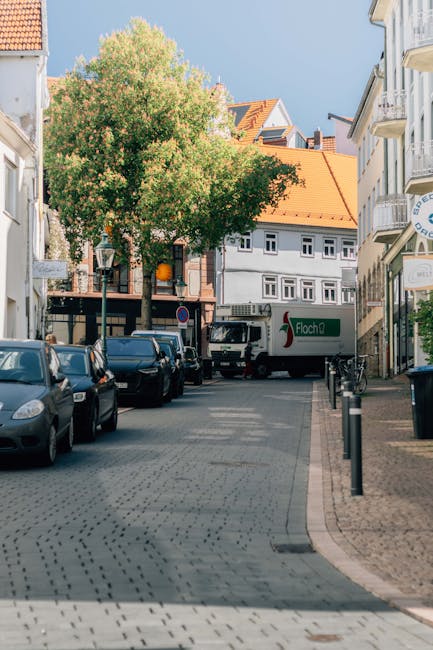 A narrow street scene in Harlington, featuring a white delivery van with green and red branding parked near a sidewalk, surrounded by various parked cars along the curb. In the background, multi-storey residential buildings with white facades and orange-tiled roofs are visible, along with a large leafy tree providing greenery to the street. The pavement is composed of grey bricks, and black bollards separate the sidewalk from the road. This setting depicts a typical urban area suitable for house removals and home relocation services, with [COMPANY_NAME] likely preparing for loading or unloading furniture and moving boxes as part of a furniture transport or packing and moving process.