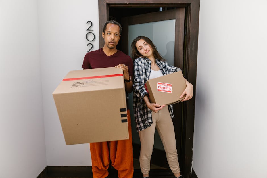 A man and a woman standing inside an entrance hallway of a residential property, preparing for a home relocation with a removal company. The man, dressed in an orange pair of trousers and a maroon T-shirt, is holding a large cardboard box with a shipping label, while the woman, wearing beige trousers and a plaid shirt, is carrying a smaller cardboard box marked with a 'Fragile' label. Both individuals appear to be waiting adjacent to a dark wooden door frame, which leads to an interior space, and are positioned near the doorway, likely during the loading process of furniture and packing materials. On the side of the wall, there are visible black house numbers, indicating the property’s address. The hallway has white walls and a black floor, and the lighting is natural and bright. This scene illustrates a typical point during packing and moving activities, which involves careful handling and transport of boxes as part of furniture transport and home relocation services provided by Man With a Van Harlington.