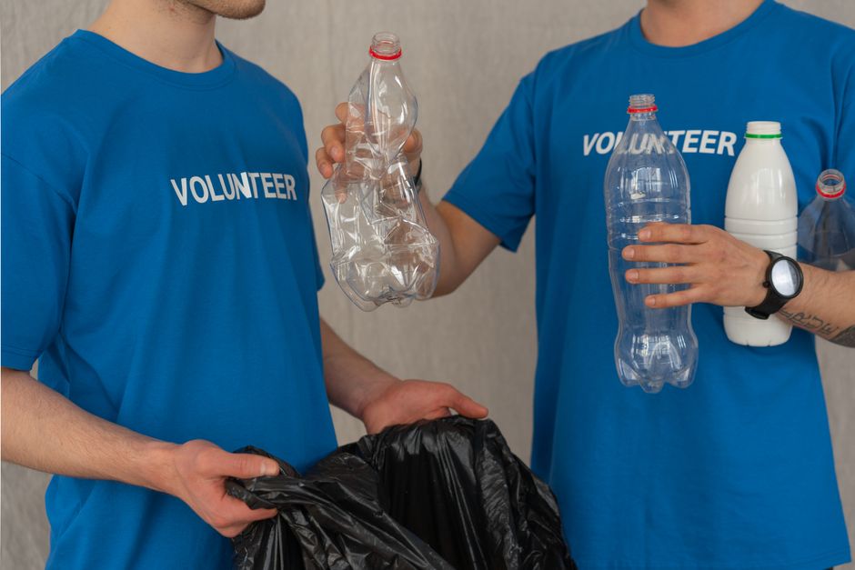 Two volunteers wearing blue T-shirts with the word 'VOLUNTEER' visible are seen during a recycling and donation event related to house removals and home relocation. One volunteer holds a crumpled plastic bottle, while the other holds a clear plastic bottle and a white plastic container, all suitable for recycling and eco-friendly disposal. The third volunteer, partially visible, is holding a black garbage bag, likely for collecting recyclable or unwanted items. They are standing indoors against a neutral wall, with soft, even lighting that highlights the various plastic objects and packing materials. The scene depicts a practical aspect of packing and moving logistics, highlighting the importance of recycling and donation options as part of an organized removal process, supported by Man With a Van Harlington.