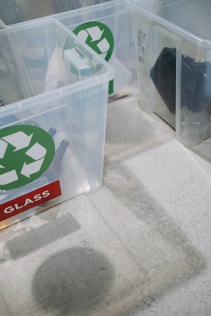 Three clear plastic recycling bins with green recycling symbols and red labels reading 'GLASS' are positioned on a light-colored carpeted floor inside a property. The bins are container-shaped with open tops, and some smaller white objects are visible inside one of them. Shadows cast on the floor suggest overhead lighting, and the bins appear to be part of a home recycling collection area, potentially for glass disposal during a house removal or moving process. The image depicts the packing and sorting stage in a home relocation, with the focus on environmentally friendly disposal options supported by Man With a Van Harlington, a removal service specializing in furniture transport and packing during moves.
