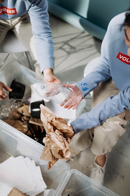 Two volunteers wearing blue T-shirts with the word 'VOLUNTEER' visible are seen during a recycling and donation event related to house removals and home relocation. One volunteer holds a crumpled plastic bottle, while the other holds a clear plastic bottle and a white plastic container, all suitable for recycling and eco-friendly disposal. The third volunteer, partially visible, is holding a black garbage bag, likely for collecting recyclable or unwanted items. They are standing indoors against a neutral wall, with soft, even lighting that highlights the various plastic objects and packing materials. The scene depicts a practical aspect of packing and moving logistics, highlighting the importance of recycling and donation options as part of an organized removal process, supported by Man With a Van Harlington.