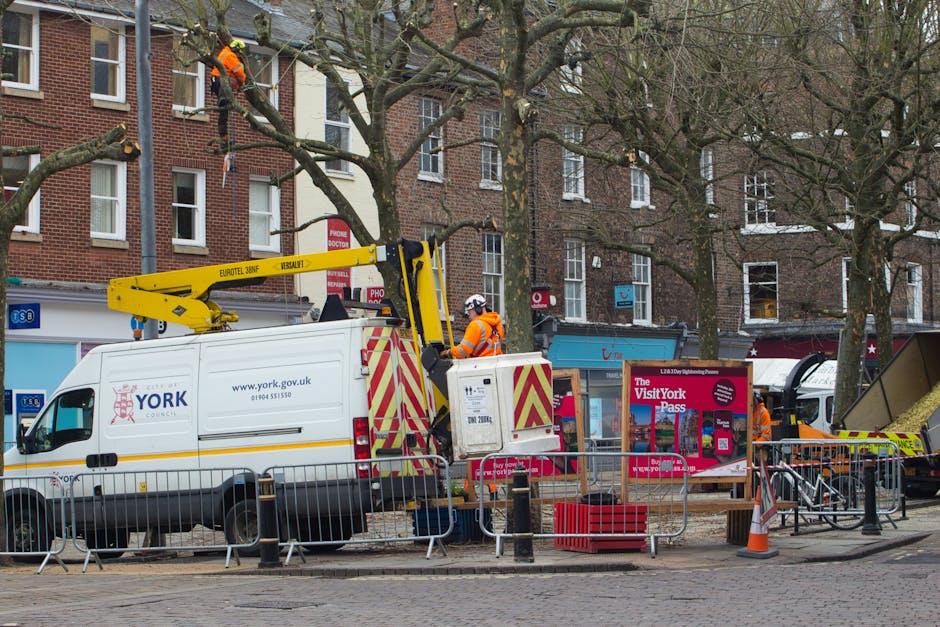 A utility van with York Council branding, equipped with a mounted cherry picker, is positioned on a street in Harlington during daytime. The cherry picker is extended upward, with a worker in high-visibility orange clothing and a safety helmet operating the equipment from the van’s elevated platform, likely engaged in tree maintenance or street work. The van is parked near a sidewalk with metal barriers set up around the working area, and a red and white signage board indicating visitor information for York Council is visible nearby. In the background, multi-story brick residential buildings, some with open windows, and leafless trees suggest a late autumn or winter setting. The scene includes other maintenance vehicles, equipment, and workers wearing orange safety vests, illustrating a typical city maintenance or utility operation related to the area’s moving or logistical arrangements, possibly connected to home relocations or street parking permits managed by [COMPANY_NAME].