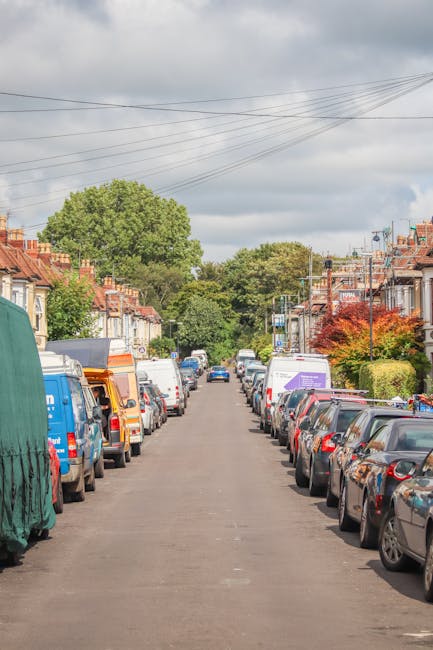 A straight residential street with parked vehicles on both sides, including various small vans, SUVs, and cars. The street is lined with terraced houses featuring brick facades, front gardens with trees and shrubs, and some houses with small front porches. Several boxes wrapped in brown cardboard and plastic are visible on the pavement near the vehicles, indicating packing for home relocation. A man dressed in casual clothing is seen lifting a box onto a trolley or moving equipment near a white van, part of a professional removals process by Man With a Van Harlington. The background shows large green trees and a partly cloudy sky, suggesting daytime with diffused natural light. Utility wires run above the street, and the environment is clean and well-maintained, typical for a residential area undergoing packing and furniture transport during a house move.