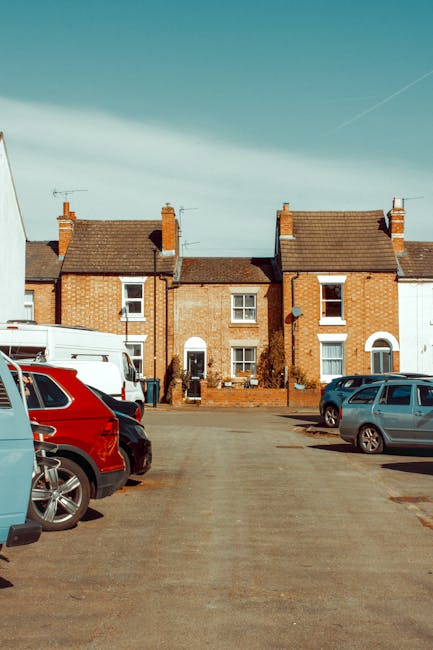A utility van with York Council branding, equipped with a mounted cherry picker, is positioned on a street in Harlington during daytime. The cherry picker is extended upward, with a worker in high-visibility orange clothing and a safety helmet operating the equipment from the van’s elevated platform, likely engaged in tree maintenance or street work. The van is parked near a sidewalk with metal barriers set up around the working area, and a red and white signage board indicating visitor information for York Council is visible nearby. In the background, multi-story brick residential buildings, some with open windows, and leafless trees suggest a late autumn or winter setting. The scene includes other maintenance vehicles, equipment, and workers wearing orange safety vests, illustrating a typical city maintenance or utility operation related to the area’s moving or logistical arrangements, possibly connected to home relocations or street parking permits managed by [COMPANY_NAME].
