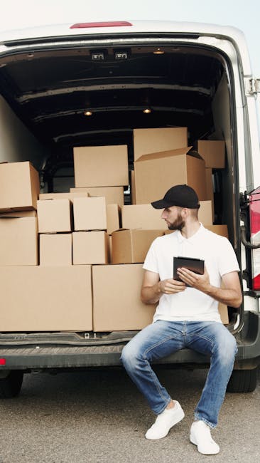 A man with a short beard, wearing a black cap, white t-shirt, and blue jeans, is sitting on the pavement beside an open van loaded with stacked cardboard boxes of various sizes, some wrapped in plastic film. The van, parked on a paved surface, is the focus of a home relocation or furniture transport process, with the interior visible showing additional boxes and packing materials. The scene occurs outdoors during daylight, and the man is holding a tablet, possibly coordinating the moving or packing activity. This image illustrates the loading process involved in house removals carried out by Man With a Van Harlington, a company specialising in moving and storage services near Hillingdon Station.