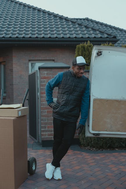 A man with a short beard, wearing a black cap, white t-shirt, and blue jeans, is sitting on the pavement beside an open van loaded with stacked cardboard boxes of various sizes, some wrapped in plastic film. The van, parked on a paved surface, is the focus of a home relocation or furniture transport process, with the interior visible showing additional boxes and packing materials. The scene occurs outdoors during daylight, and the man is holding a tablet, possibly coordinating the moving or packing activity. This image illustrates the loading process involved in house removals carried out by Man With a Van Harlington, a company specialising in moving and storage services near Hillingdon Station.