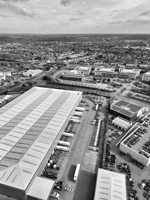 A black-and-white aerial photograph showing a large industrial warehouse with a metal roof and multiple loading bays on the left side of the image. Several delivery trucks are parked in designated areas along the loading bays, with some trucks positioned at the entrances, possibly being loaded or unloaded. To the right, there are several smaller commercial or office buildings with flat roofs, surrounded by a parking lot filled with numerous parked cars. The parking lot includes marked spaces, with some vehicles parked parallel to the buildings and others at angles. Behind the warehouse and office complex, an open space with pathways and additional parking facilities is visible, along with a network of roads leading to and from the site. In the background, residential and commercial areas extend towards the horizon, with a mix of buildings, green spaces, and roadways, under a partly cloudy sky. This scene illustrates elements of logistics and transportation relevant to house removals and furniture transport services, highlighting the loading process and planning involved in moving projects, as managed by [COMPANY_NAME].