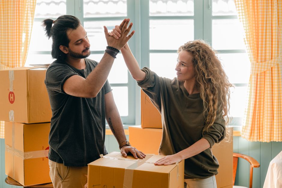 A man and woman inside a room during a home relocation, engaging in a celebratory gesture with their hands. The man, with dark hair tied back, is wearing a dark t-shirt and has a bracelet on his wrist