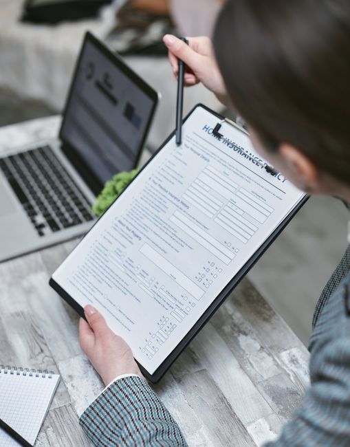 A person reviewing a detailed moving checklist or insurance document on a clipboard during a home relocation process, with a laptop open nearby displaying related information. The individual is seated at a weathered wooden table inside a property, surrounded by packing materials such as cardboard boxes, plastic wrapping, and foam padding. The scene captures the preparation phase of a house move, with visible objects including boxes stacked in the background, some wrapped furniture parts, and the person's hand holding a pen as they point to specific details on the document. Natural lighting illuminates the workspace, emphasizing the careful planning involved in furniture transport and packing logistics, consistent with services offered by Man With a Van Harlington for efficient removals and insurance explanations.