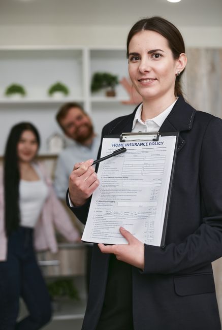 A person reviewing a detailed moving checklist or insurance document on a clipboard during a home relocation process, with a laptop open nearby displaying related information. The individual is seated at a weathered wooden table inside a property, surrounded by packing materials such as cardboard boxes, plastic wrapping, and foam padding. The scene captures the preparation phase of a house move, with visible objects including boxes stacked in the background, some wrapped furniture parts, and the person's hand holding a pen as they point to specific details on the document. Natural lighting illuminates the workspace, emphasizing the careful planning involved in furniture transport and packing logistics, consistent with services offered by Man With a Van Harlington for efficient removals and insurance explanations.