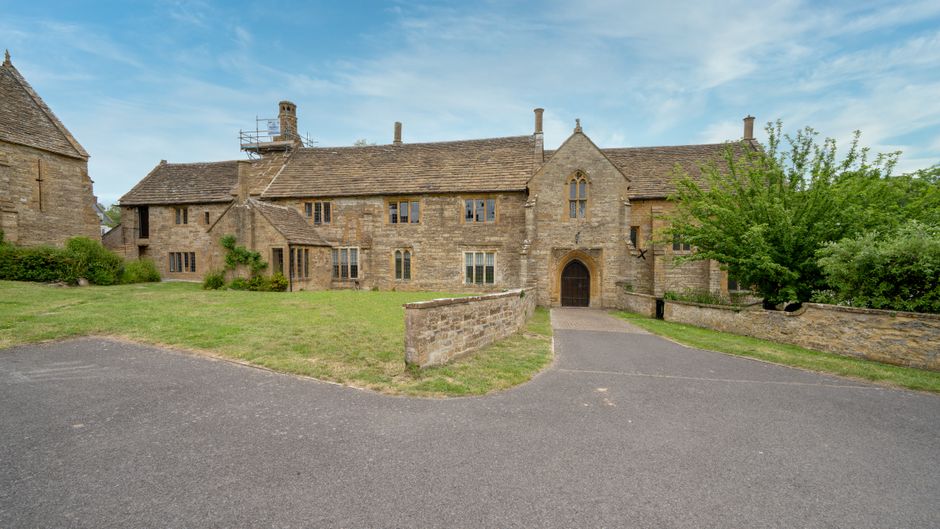A large, historic stone building with a pitched roof and multiple chimneys, situated on a spacious property with a paved driveway leading up to an arched wooden door. The building features several win