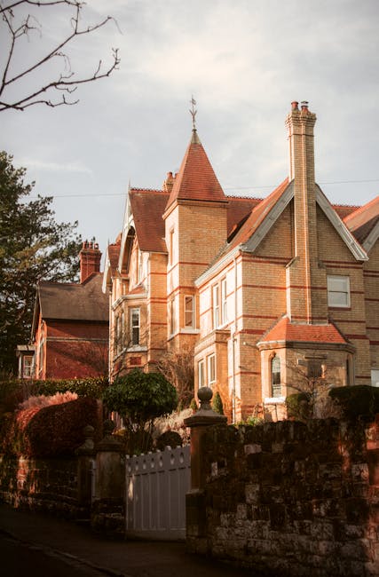 A large, historic stone building with a pitched roof and multiple chimneys, situated on a spacious property with a paved driveway leading up to an arched wooden door. The building features several win