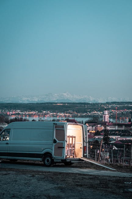 A large white moving van parked on a paved area with its side door open, revealing a wooden staircase and packing materials inside, including cardboard boxes and fabric blankets used for protecting furniture during a home relocation. The van is positioned on an incline with a ramp extended on the ground, facilitating loading and unloading. In the background, there is a panoramic view of a cityscape with industrial buildings, residential areas, and distant snow-capped mountains under an overcast sky. The scene reflects the process of furniture transport and packing involved in professional removals services, as provided by Man With a Van Harlington, situated within an urban environment suitable for house moves and logistics coordination.