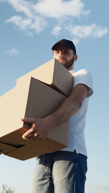 A man with a beard and wearing a black cap, white t-shirt, and jeans is standing outdoors on a clear day with a blue sky and a few scattered clouds. He is holding three cardboard boxes, stacked one on top of the other, securely against his chest with both arms, preparing for a home relocation. The boxes are plain brown with no visible labels or markings. Behind him, a paved area or driveway is visible, suggesting a residential property or moving site. The lighting indicates natural sunlight, creating a well-lit scene suitable for documenting furniture transport or loading activities associated with same-day removals. This image aligns with professional removal services offered by Man With a Van Harlington, emphasizing packing, loading, and transport processes involved in house moves or last-minute bookings.