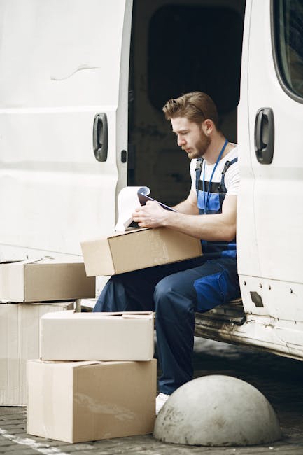 A man dressed in casual work clothing, sitting on the edge of a white van with the side door open, engaged in reading or reviewing papers while handling a cardboard box during a home relocation. Several unpacked cardboard boxes of various sizes are placed on the ground nearby, some positioned on top of each other. The van is parked on a paved surface, adjacent to a concrete bollard, with outdoor lighting illuminating the scene. This image depicts the loading or packing process associated with furniture transport and house removals, illustrating professional moving logistics. The environment appears to be an exterior setting, possibly in a driveway or parking area, with the focus on the man working on packing or organizing items in preparation for a moving service provided by Man With a Van Harlington, supporting the context of same day removals and last-minute bookings.