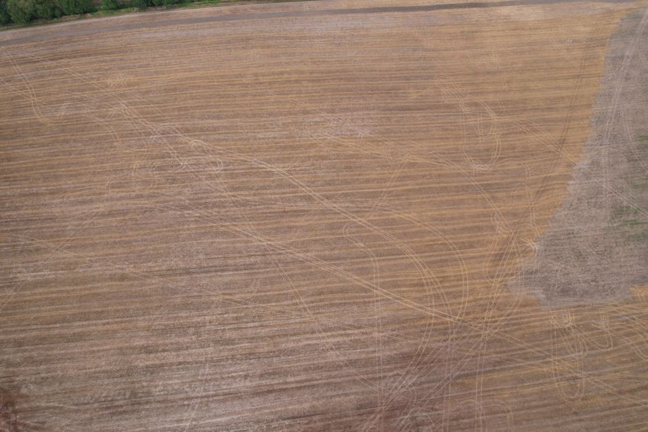 An aerial view of a large area of land with visible tire marks and paths, featuring a mixture of brown and tan soil with some patches of grass on the right side. The tire tracks, likely created during moving or transportation activities, cross diagonally and in various directions across the open ground. The terrain appears flat, with no buildings or structures present. This image may depict the environment surrounding a property where house removals and furniture transport are taking place, highlighting the challenges of home relocation involving outdoor access issues. The presence of vehicle tracks suggests recent activity related to loading or unloading furniture and boxes, consistent with the services provided by Man With a Van Harlington, especially when dealing with stair access problems during a home move.